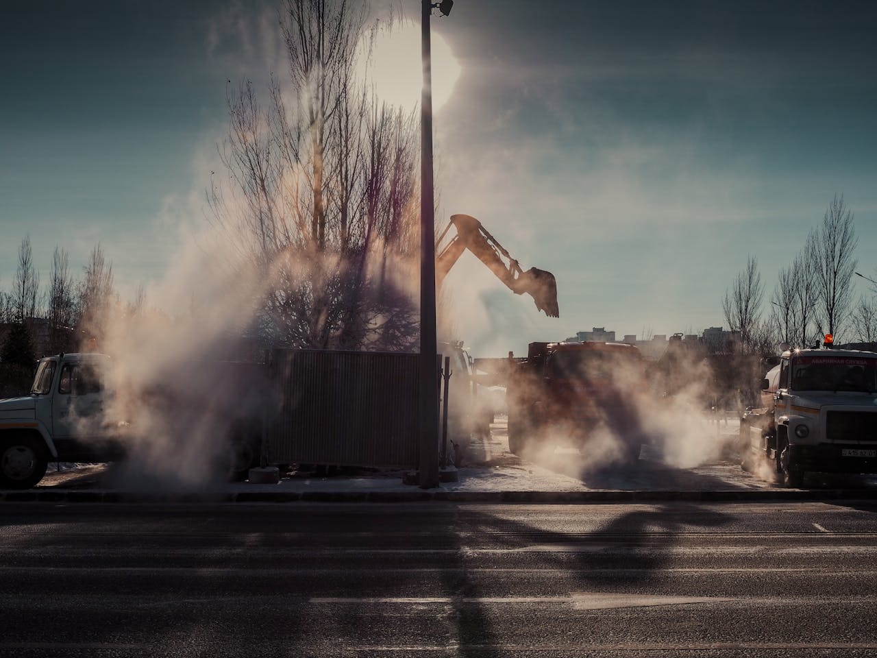 A dramatic scene of construction work on a city street with machinery and steam at sunrise.