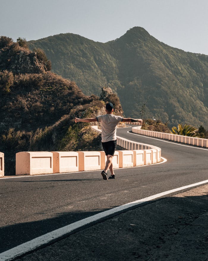 A person with arms wide open walks along a winding mountain road in Tenerife, Spain, surrounded by lush hills.