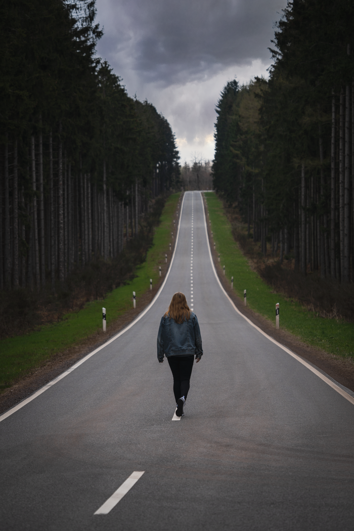 A lone woman walks on a serene forest road beneath a moody, cloudy sky. Captured from behind.