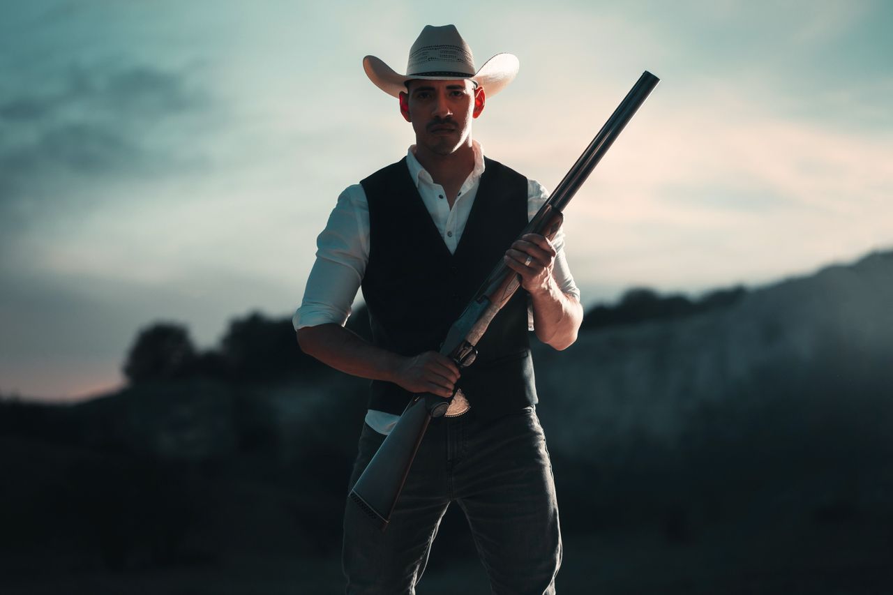 about-us A cowboy in traditional attire holding a shotgun, standing outdoors in San Antonio, Texas.