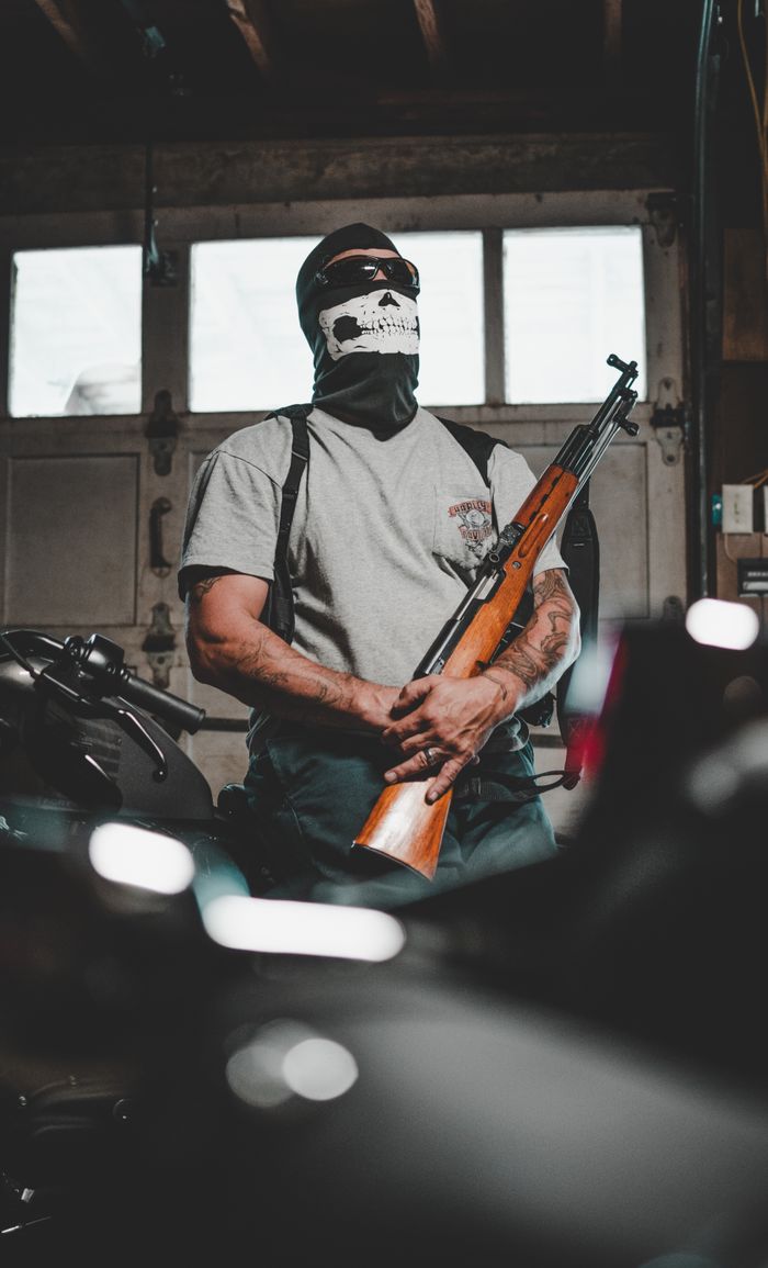heros-img A masked person stands in a garage holding a rifle, surrounded by motorcycles.