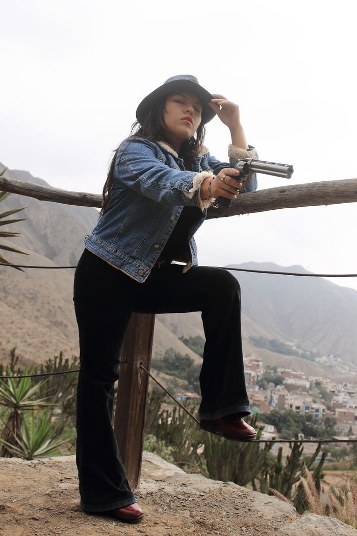 Stylish cowgirl in denim and cowboy hat with a revolver, set against the scenic mountains of Lima, Peru.